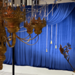 Prabangi mėlyna foto sienelė su audiniu, dekoruota auksinėmis virvelėmis ir kutais, sukuria elegantišką atmosferą su apšvietimo efektais.